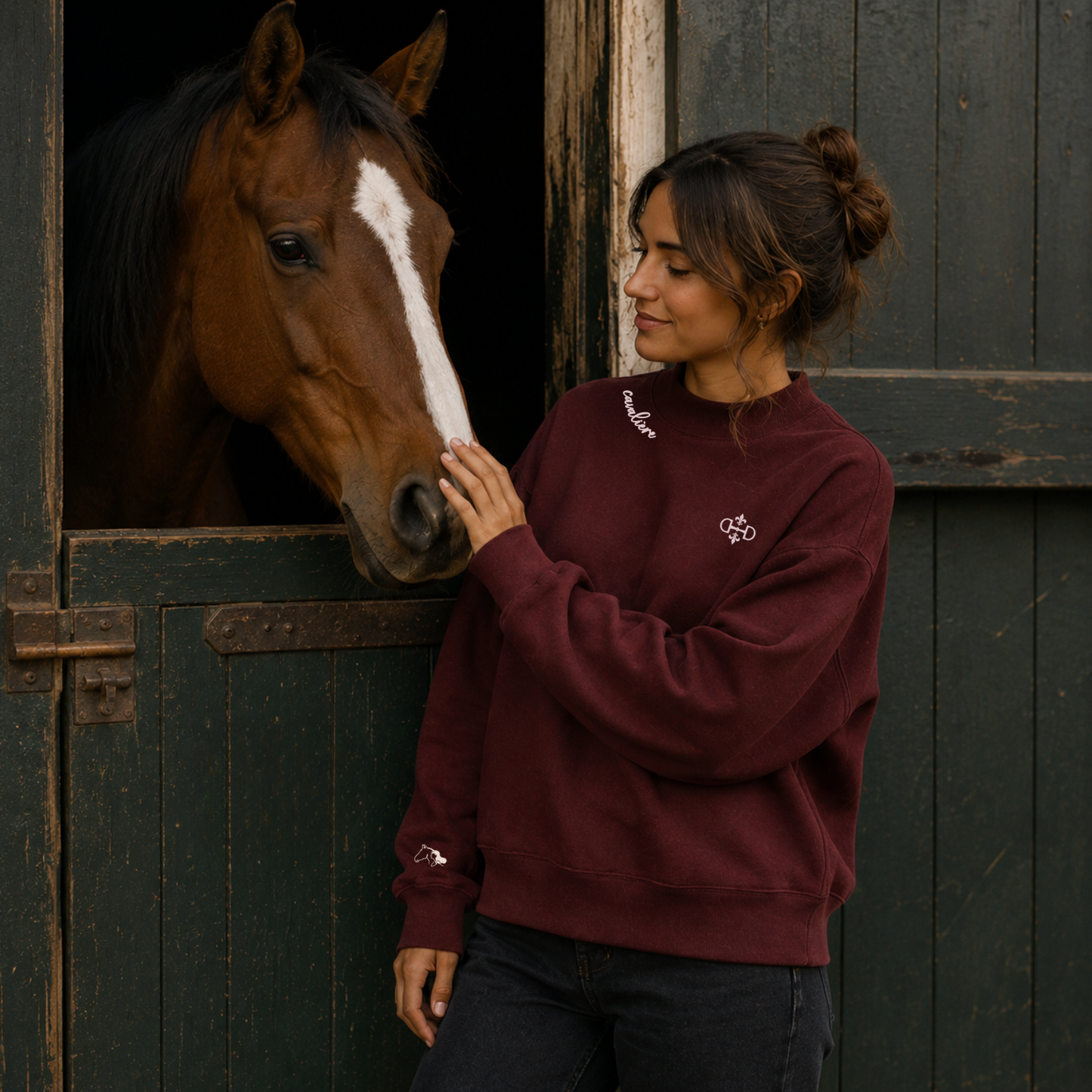 A woman in a burgundy mock neck crewneck sweatshirt with blush embroidered logos stands next to a brown horse with a white blaze, touching its face inside a dark barn stable.