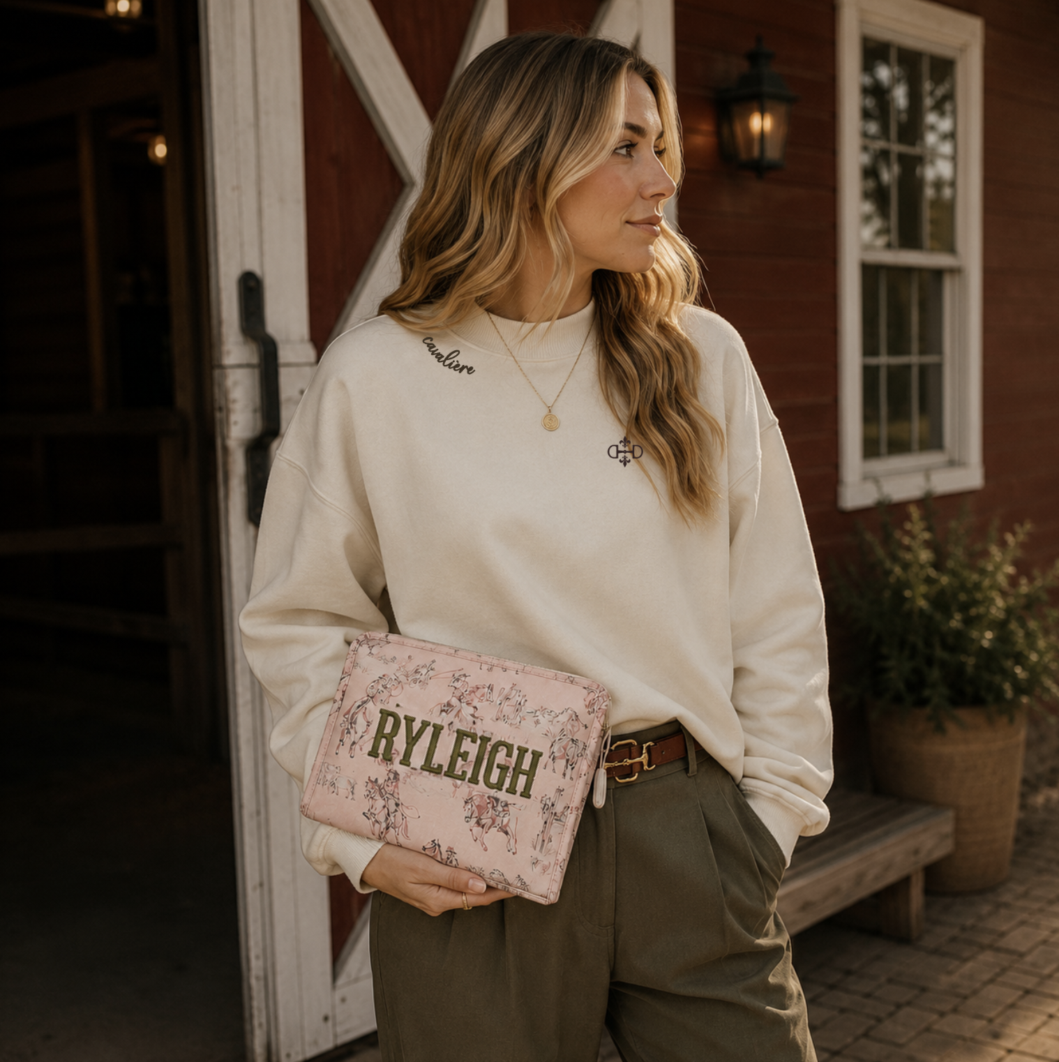 A woman stands in front of a rustic red barn doorway wearing a cream mock neck crewneck with chocolate embroidery, holding a light pink cowgirl toile clutch bag with the name 'RYLEIGH' embroidered on the front.