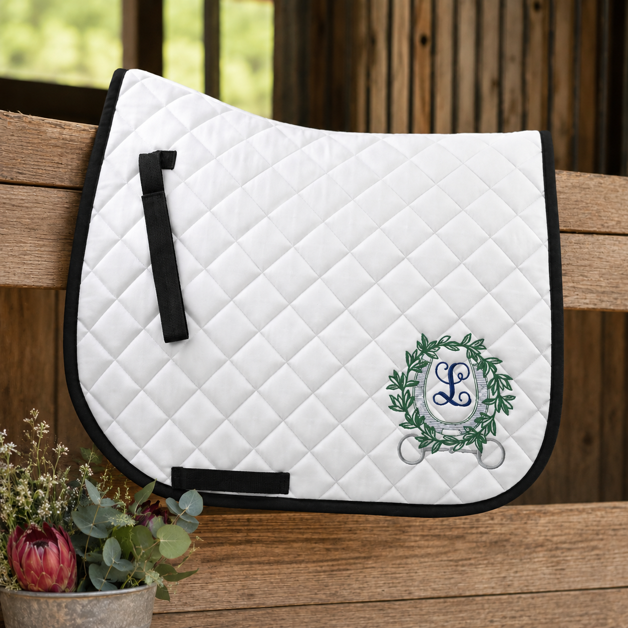 A white quilted equestrian saddle pad with black trim and straps, featuring a green wreath monogram with a blue 'L' on the front, resting on a wooden bench.