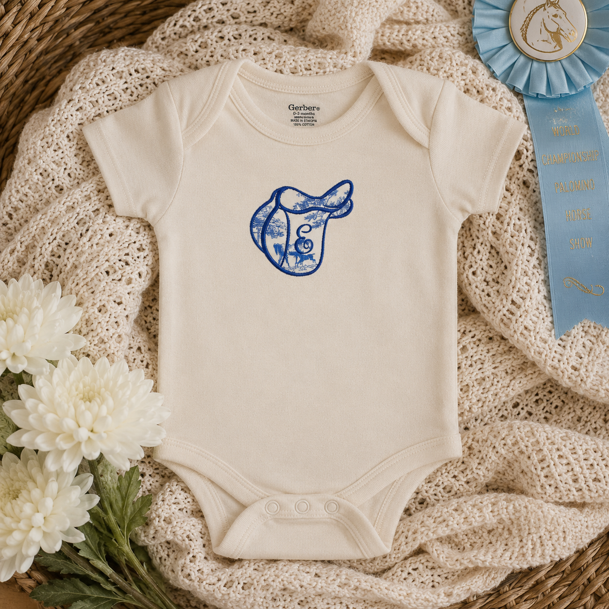 An off-white short-sleeve baby bodysuit with a blue horse saddle graphic on the chest, styled on a beige crochet blanket with white flowers and a blue ribbon in a wicker basket.