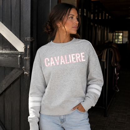 A woman wearing a light gray heather crewneck sweatshirt with embroidered  'CAVALIERE' applique in a pink horse toile fabric across the chest and white stripes on the sleeves, standing in a dark barn interior.