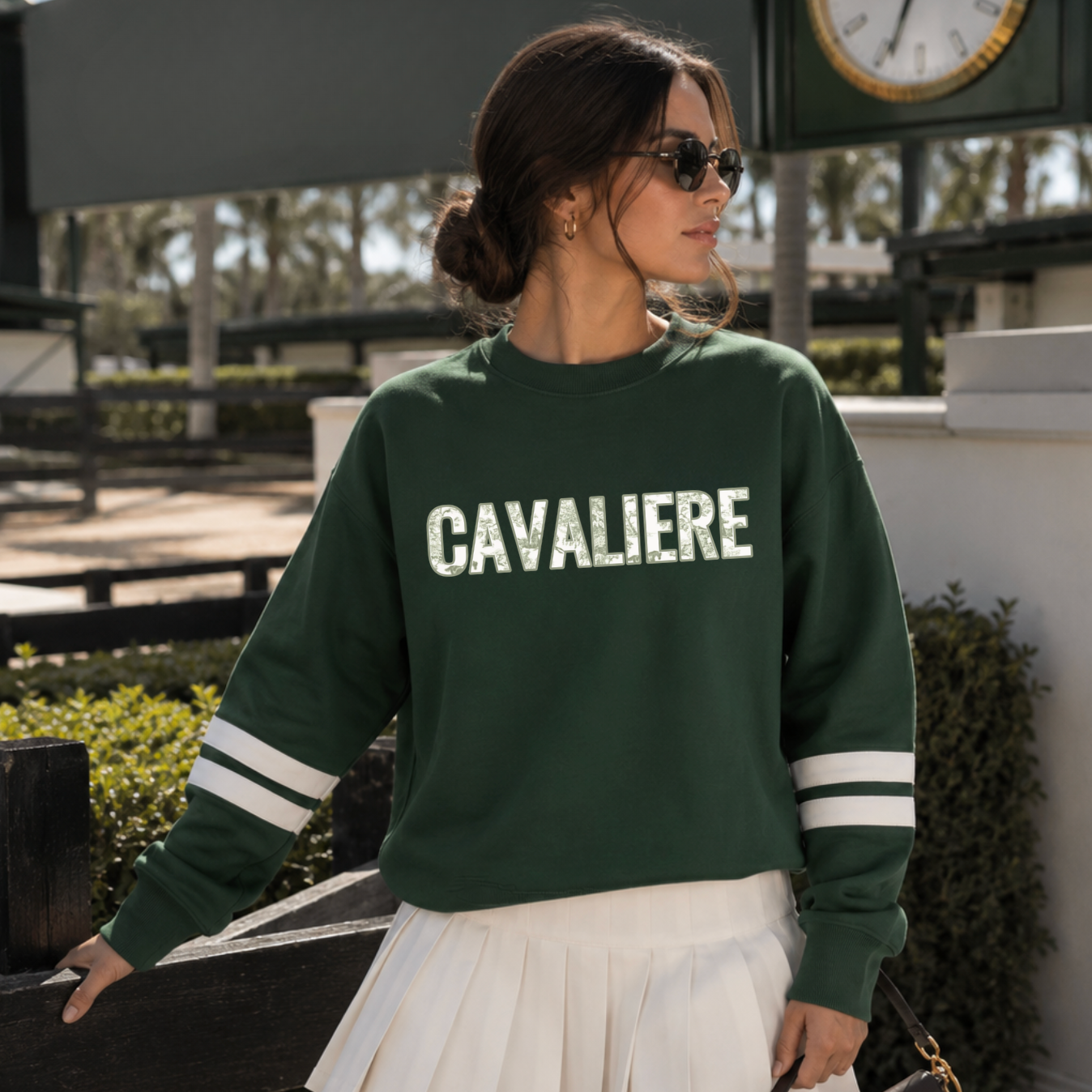 A woman wearing a dark green crewneck sweatshirt with embroidered 'CAVALIERE' applique in a green horse toile fabric across the chest and white stripes on the sleeves, paired with a white pleated skirt, standing outdoors near an equestrian-themed building.