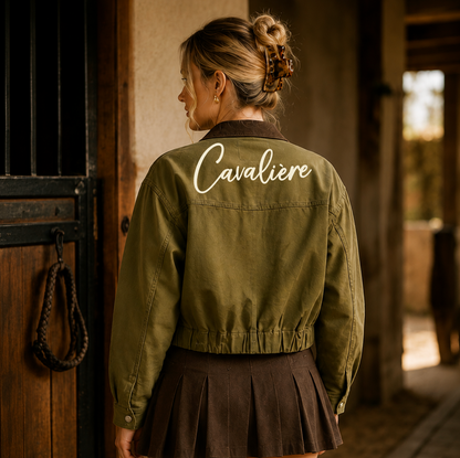 A woman stands with her back to the camera wearing an olive green cropped jacket with 'Cavaliere' embroidered in white script across the upper back, paired with a dark brown pleated skirt, in a rustic indoor stable setting.