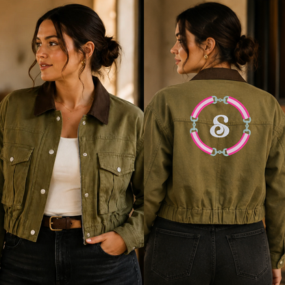 A woman modeling an olive green cropped utility jacket, shown from both the front and back. The jacket features a dark brown collar, silver snap buttons, and embroidery on the back with a pink neon ribbon circle, silver snaffle bits, and a white monogram 'S'.