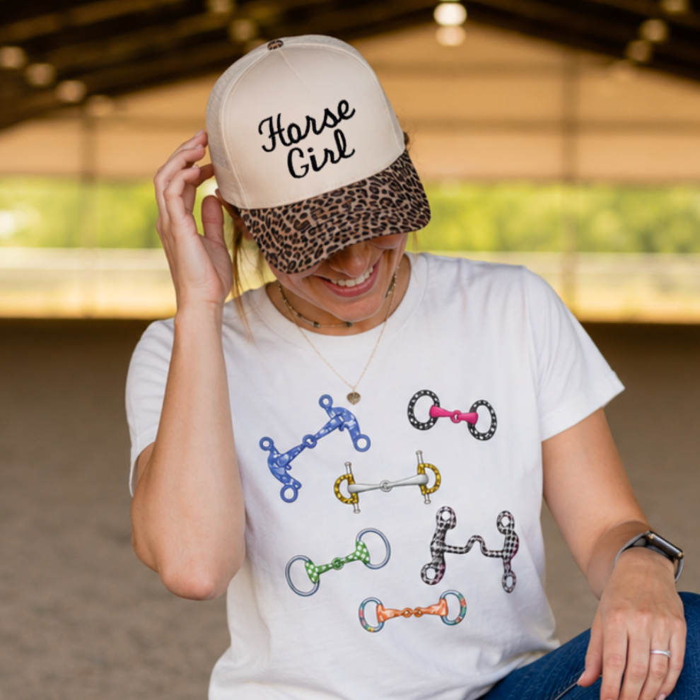 A woman kneels in an indoor horse arena wearing a white graphic t-shirt with horse bit designs, blue jeans, and a beige 'Horse Girl' cap.