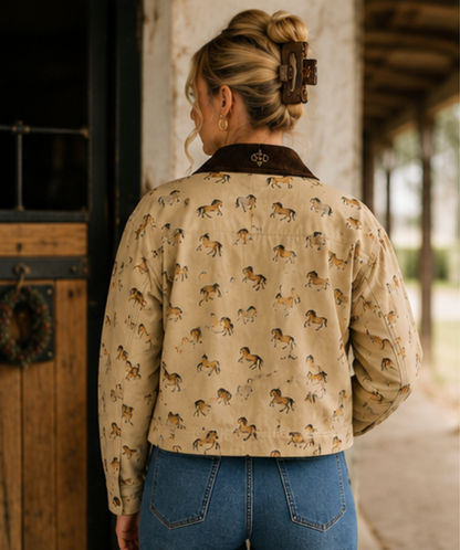 A woman viewed from behind wearing a cropped beige jacket with an allover horse print and a dark brown collar, paired with blue jeans in a rustic covered porch setting.