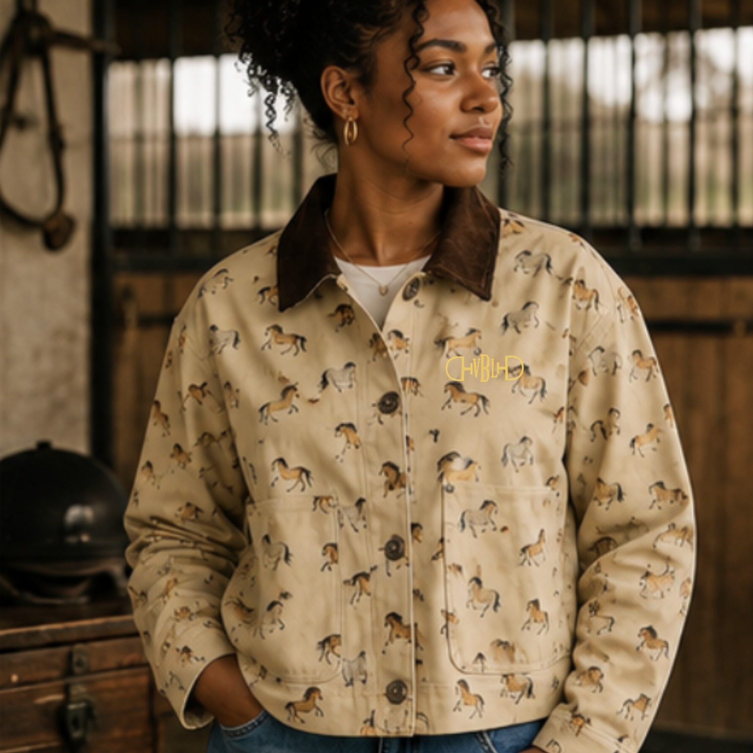 A woman wearing a beige utility-style jacket with an allover horse print and a dark brown collar and custom monogram embroidery, standing in a rustic indoor setting with horse stalls in the background.