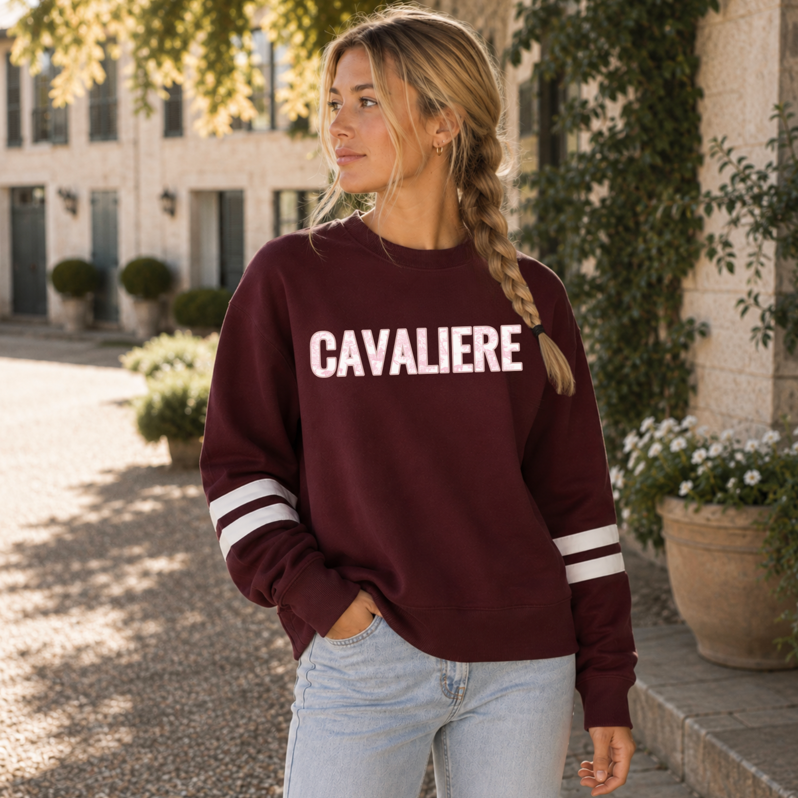 A woman wearing a maroon crewneck sweatshirt with embroidered 'CAVALIERE' applique in a white and pink horse toile fabric across the chest and white stripes on the sleeves, paired with light blue jeans, standing outdoors in front of a stone building.