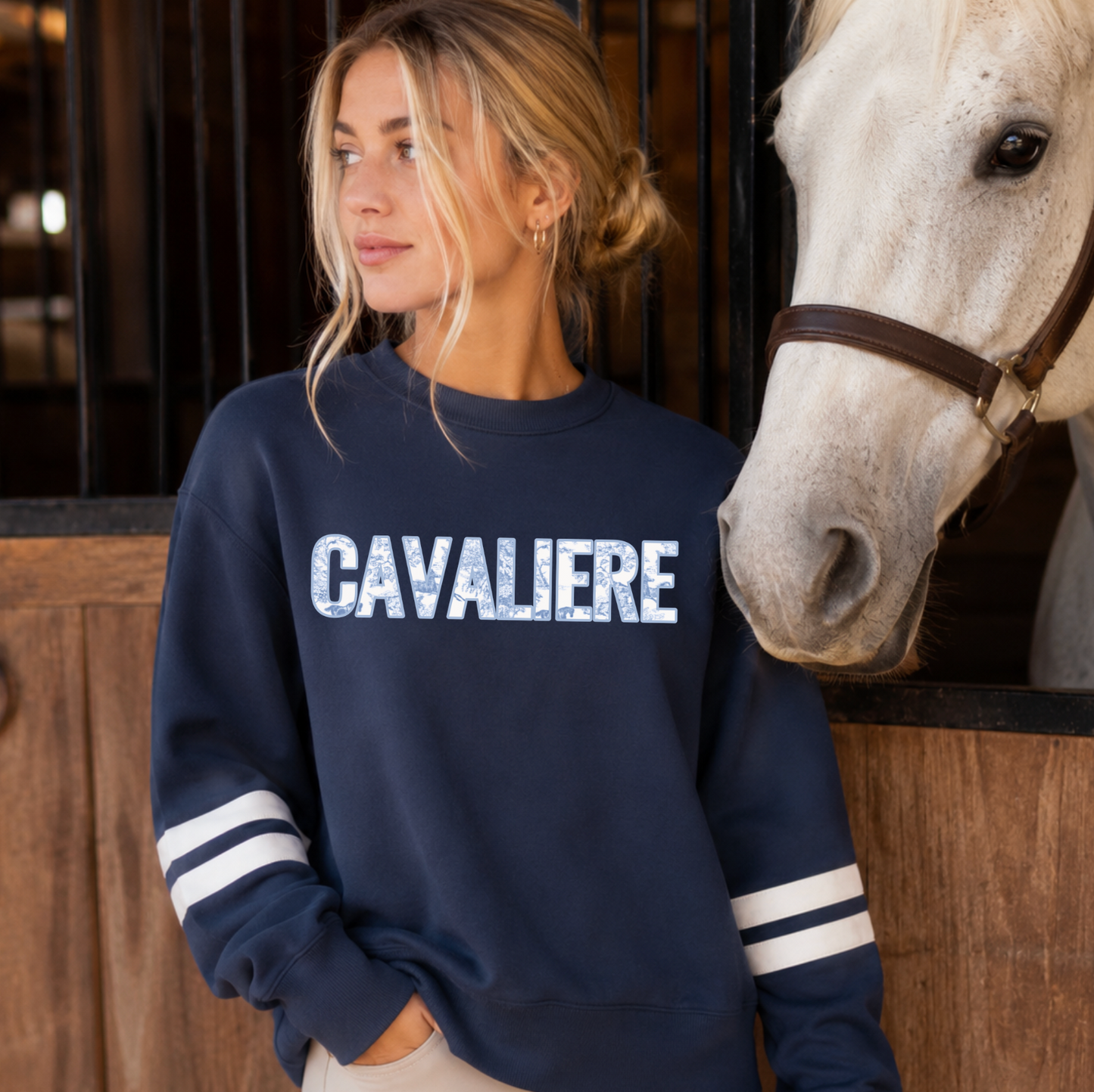 A woman wearing a navy blue varsity style sweatshirt with embroidered 'CAVALIERE' applique across the chest in a white and blue horse toile fabric, standing next to a white horse in a stable. The sweatshirt has white stripes on the sleeves.