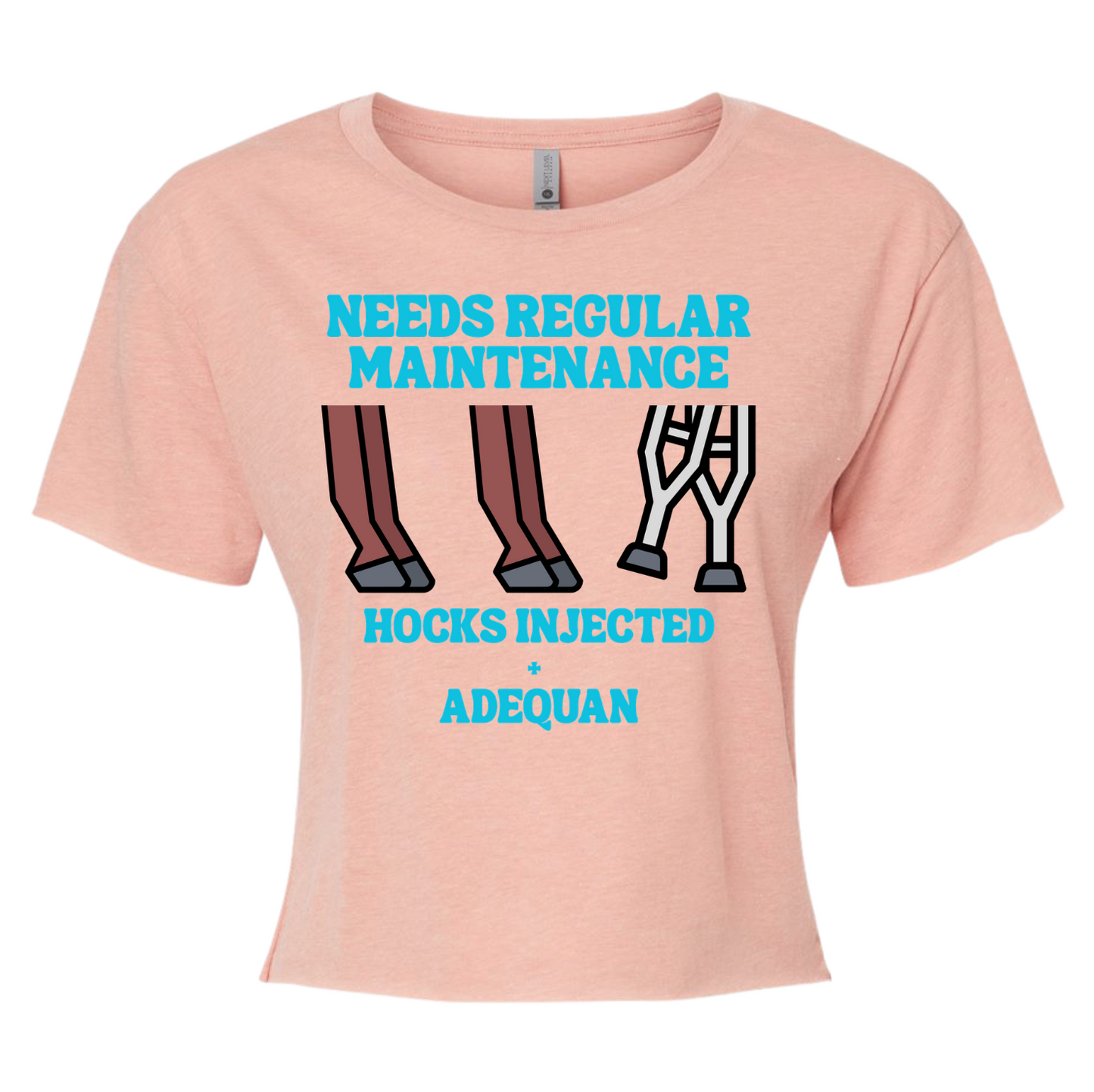 A light peach-pink short-sleeve crop top t-shirt with a graphic print. The graphic features the text 'NEEDS REGULAR MAINTENANCE' and 'HOCKS INJECTED + ADEQUAN' in bright cyan blue, along with illustrations of horse legs (one brown, one white with casters).