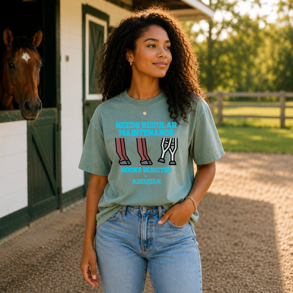 A woman stands outdoors in front of a horse stable wearing a sage green graphic t-shirt with a humorous horse care slogan and leg illustration. A horse is visible in the barn doorway on the left, and a grassy field with a fence is in the background.