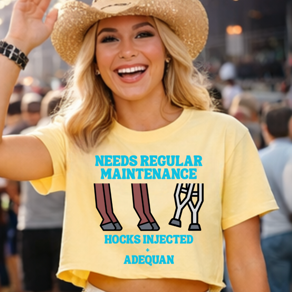 A smiling blonde woman at an outdoor event wears a straw cowboy hat, a bright yellow graphic T-shirt that reads 'NEEDS REGULAR MAINTENANCE' and 'HOCKS INJECTED + ADEQUAN' with illustrated legs, and crutches.