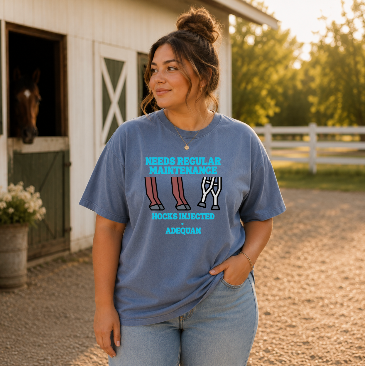 A woman stands outdoors in front of a barn wearing a blue graphic T-shirt with a humorous farm-themed slogan and illustrations of legs and crutches on the front. The background shows a horse in a barn stall, trees, and a fence.