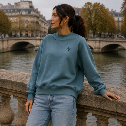 A woman wearing a sea blue mock neck crewneck sweatshirt and light wash jeans stands on a stone bridge with a river and Parisian-style buildings in the background.