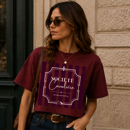 A woman wearing a burgundy short-sleeve graphic T-shirt with a purple and white 'Société Cavalière' print, paired with dark blue jeans and sunglasses, standing outdoors against a stone wall.