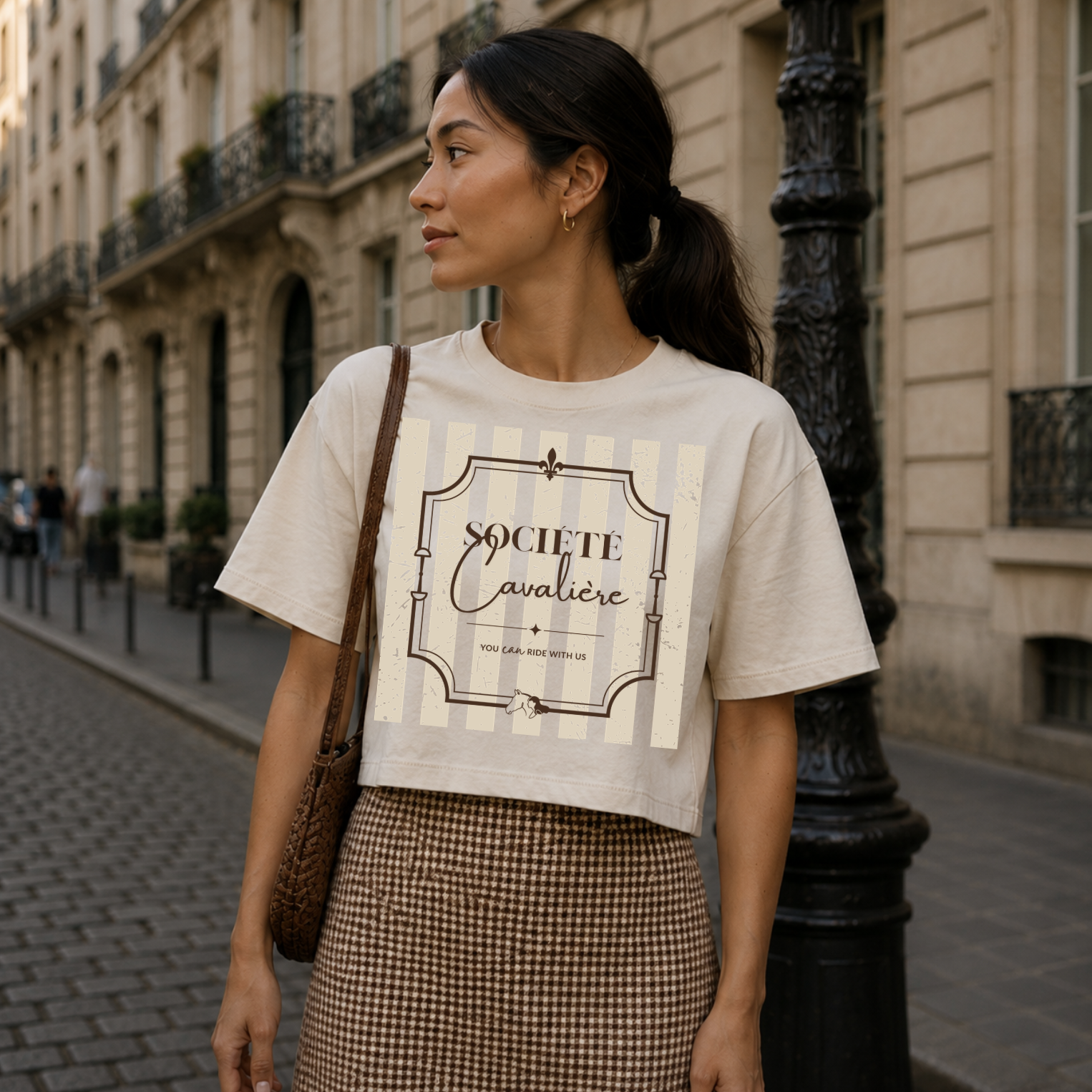 A woman stands on a city street wearing a cropped off-white graphic T-shirt with vertical cream stripes and a large decorative text print reading 'SOCIETE Cavalieri' with smaller script beneath. She is also wearing a brown-and-white houndstooth skirt and carrying a brown shoulder bag.