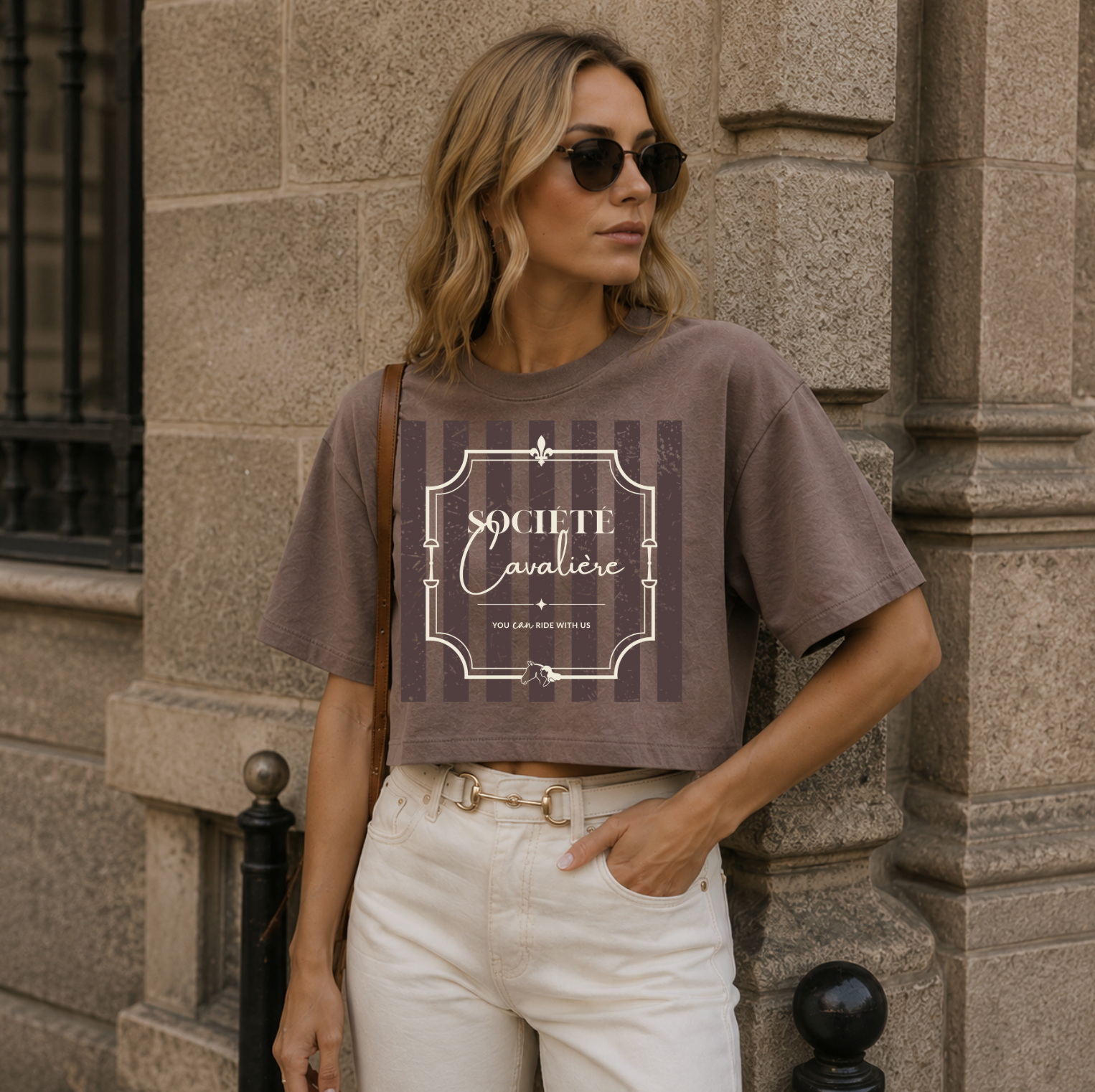 A woman wearing a brown cropped graphic t-shirt with 'Societe Cavaliere' printed on the front, paired with white jeans, standing against a stone building.