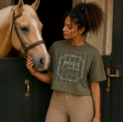 A woman wearing an olive green graphic t-shirt and beige pants stands next to a light brown horse in front of dark wooden horse stalls. The t-shirt features a graphic with the text 'SOCIETE Cavaliere'.