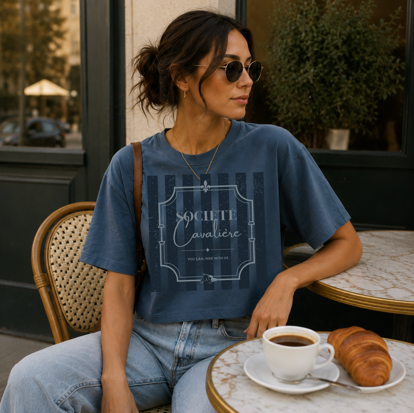 A woman wearing a blue graphic t-shirt and light-wash jeans sits at an outdoor cafe table with a cup of coffee and a croissant. The t-shirt features a faded vertical stripe pattern with a central crest graphic that reads 'SOCIETE Navale'.