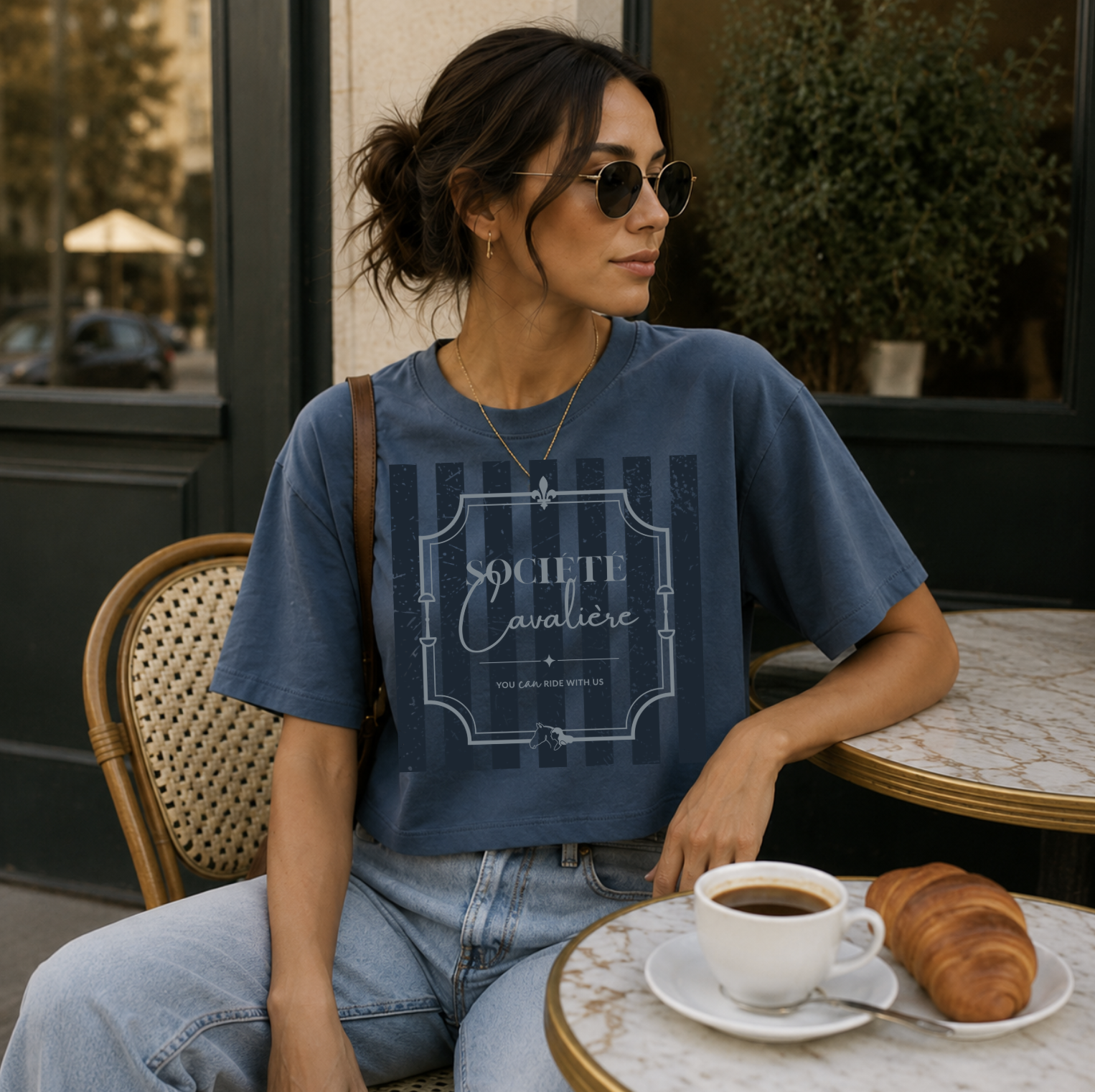 A woman wearing a blue graphic t-shirt and light-wash jeans sits at an outdoor cafe table with a cup of coffee and a croissant. The t-shirt features a faded vertical stripe pattern with a central crest graphic that reads 'SOCIETE Navale'.
