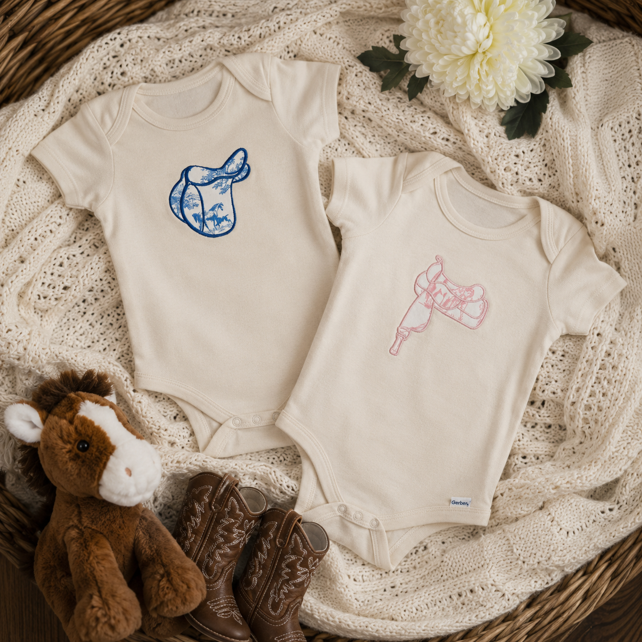Two cream-colored short-sleeve baby bodysuits laid flat on a knitted cream blanket, styled with a brown plush horse, brown cowboy boots, and a white flower. The bodysuit on the left has a blue embroidered English saddle design, and the bodysuit on the right has a pink embroidered Western saddle design.