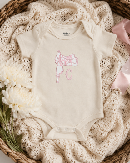 A cream baby bodysuit with a pink embroidered saddle and bow design, styled in a flat lay on a beige crochet blanket inside a wicker basket with white flowers and a pink ribbon.