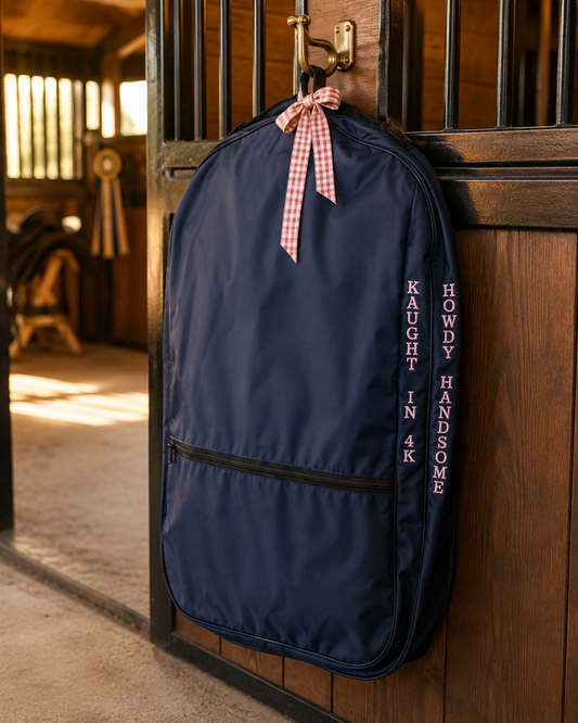 A navy blue equestrian gear bag hanging on a wooden stable door, tied with a pink and white gingham ribbon. The bag features a front zippered pocket and vertical text reading 'KAUGHT IN 4K' and 'HOWDY HANDSOME'.