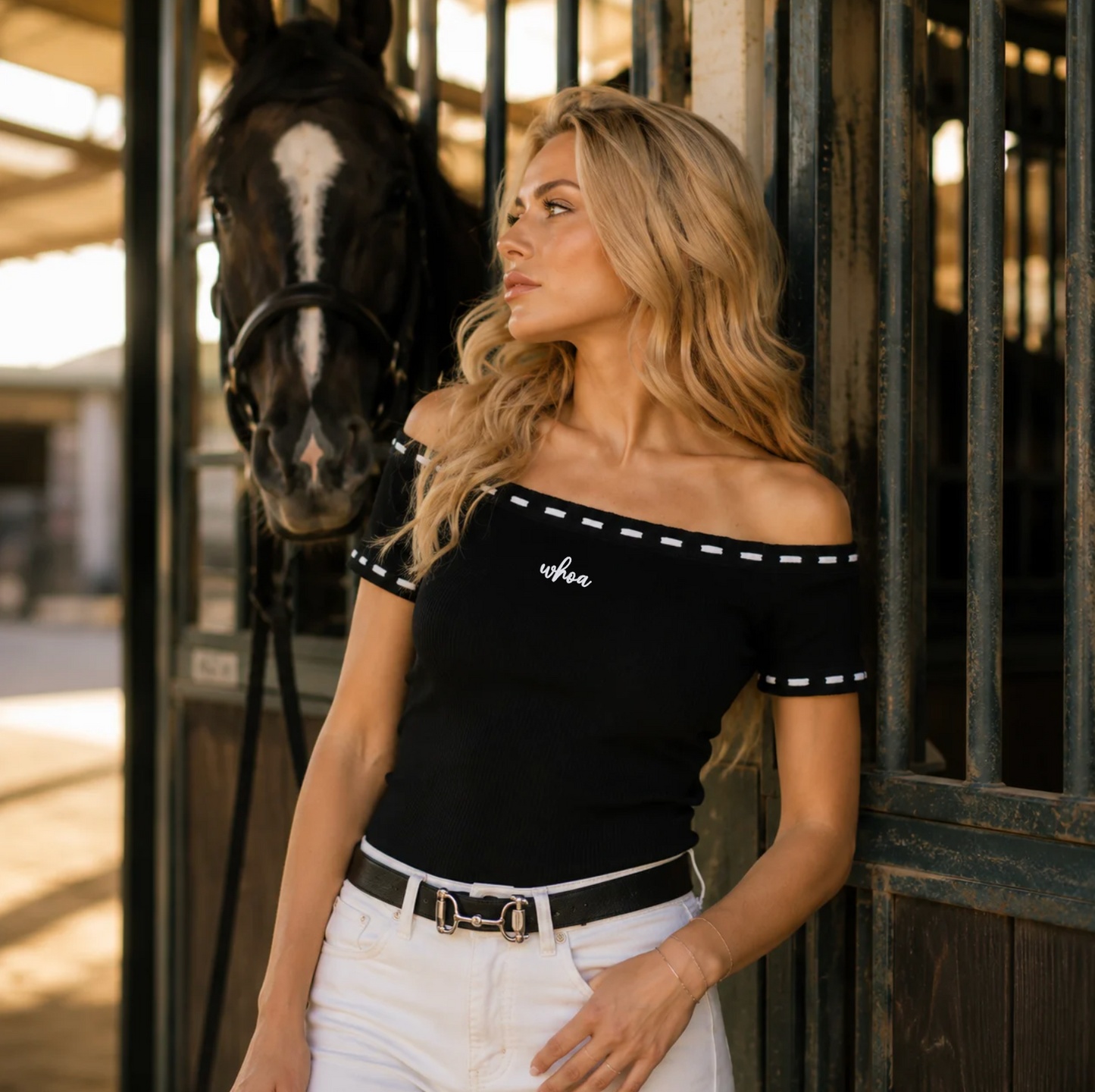 Woman wearing black off the shoulder top with white whip stitching and the word "whoa" embroidered in white on the front standing in front of a barn with a horse behind her.