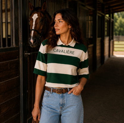 A woman wearing a green and cream striped polo shirt with 'CAVALIÈRE' printed on the chest, standing in a stable corridor with a horse behind her.