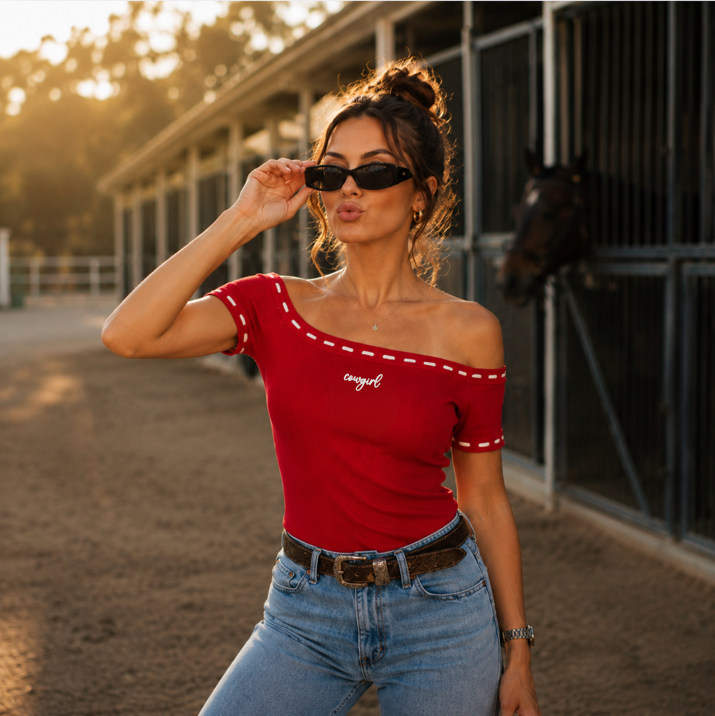 Woman wearing red off the shoulder top with white whip stitching and the word "cowgirl" embroidered in white on the front standing in front of a barn.