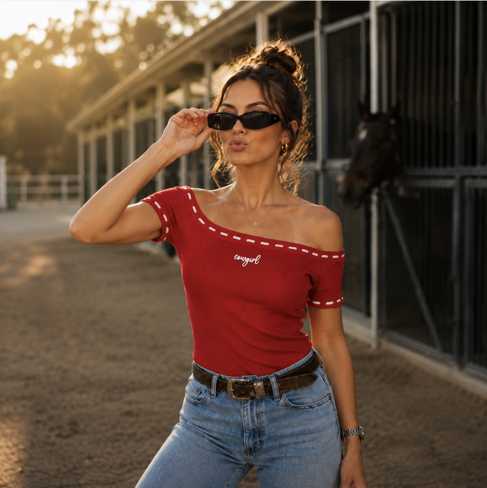 Woman wearing red off the shoulder top with white whip stitching and the word "cowgirl" embroidered in white on the front standing in front of a barn.