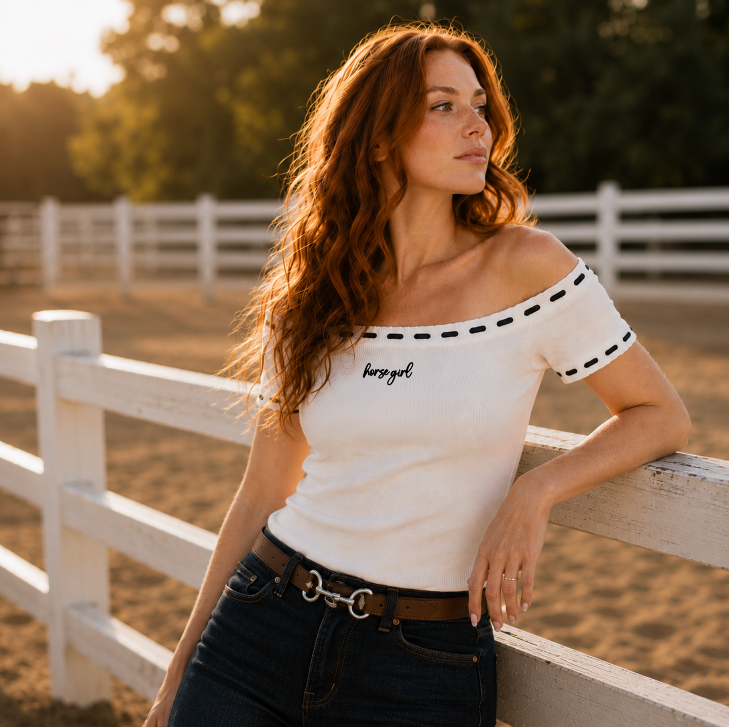 A woman with long wavy red hair leans against a white wooden fence in a sunlit outdoor setting, wearing an off-the-shoulder white fitted top with black dashed trim and the words 'horse girl' embroidered in black script on the chest, paired with dark jeans and a brown belt.